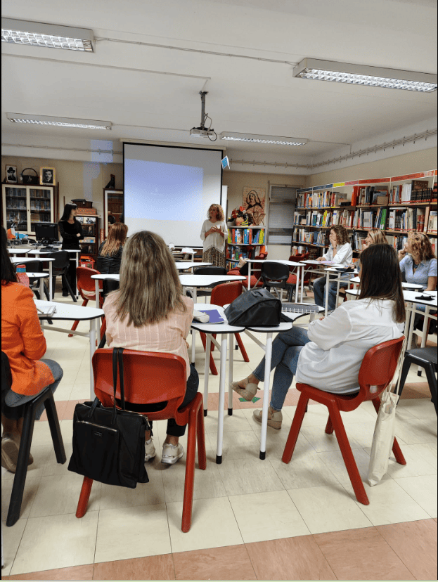 Uma mulher está em pé a falar para várias pessoas sentadas em cadeiras vermelhas, à volta de mesas brancas. Estão numa sala com muitas estantes cheias de livros. Na parede há uma imagem religiosa. Atrás da mulher há um ecrã grande com fundo azul. As pessoas têm mochilas e cadernos, como se estivessem numa aula ou reunião. A sala está bem iluminada e tudo parece calmo e organizado.