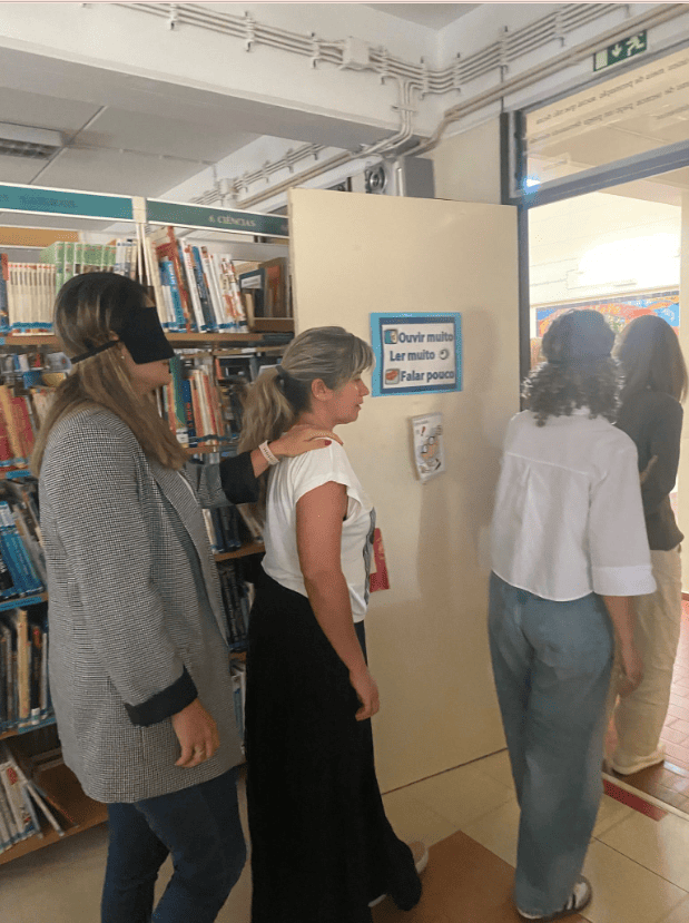 Um grupo de pessoas está a andar em fila dentro de uma sala com livros. Cada pessoa tem a mão no ombro da pessoa da frente. Uma delas está de olhos tapados. Parece uma atividade de confiança ou de aprendizagem. Na parede há uma frase que diz: “Ouvir muito, ler muito, falar pouco.”