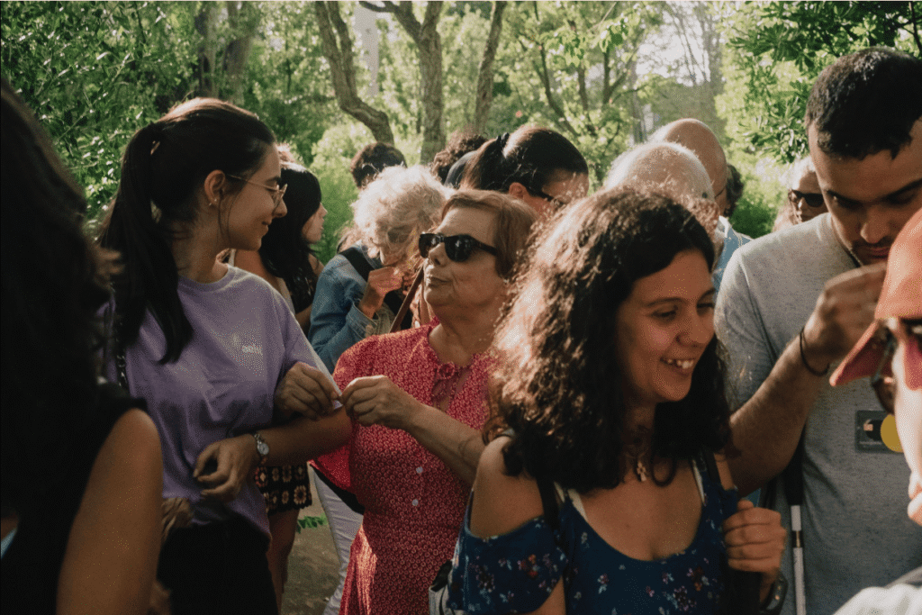 A imagem mostra um grupo de pessoas a caminhar juntas num espaço ao ar livre, rodeado de árvores e vegetação. Há pessoas de várias idades, algumas a sorrir e outras a conversar. No centro, uma mulher mais velha segura o braço de uma jovem para se orientar. O ambiente parece calmo e agradável, com luz natural a passar pelas árvores.