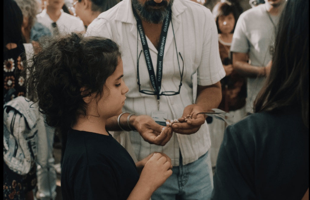 A imagem mostra uma criança a tocar num pequeno objeto que um adulto lhe está a mostrar. O adulto está ligeiramente inclinado para a criança e segura o objeto com as duas mãos, guiando-a. À volta deles há várias pessoas, como se estivessem numa atividade ou evento em grupo. O ambiente parece tranquilo e acolhedor.