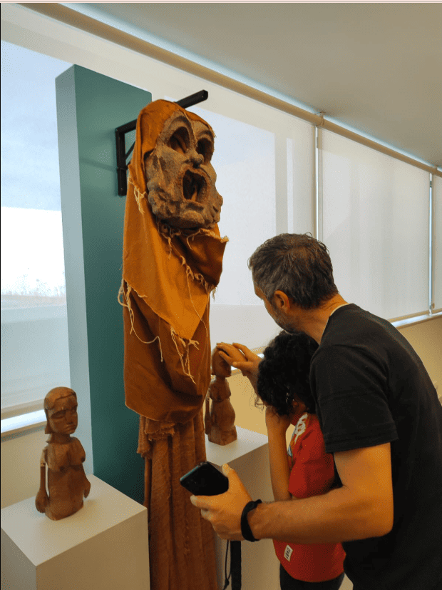A imagem mostra um homem e uma criança, vistos de costas, a tocar numa figura de madeira exposta num museu. A figura é alta, tem uma cabeça esculpida com expressão exagerada e está coberta por um tecido castanho. O homem ajuda a criança a explorar a peça com as mãos. Ao lado, em pedestais brancos, estão duas esculturas mais pequenas também feitas em madeira.
