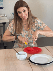 Uma pessoa está sentada à mesa e está a cortar um objeto vermelho com faca e garfo. Esse objeto está num prato vermelho. A pessoa tem um vestido com flores e várias pulseiras. Na mesa há também uma taça branca com uma colher, um copo de plástico e pratos empilhados.