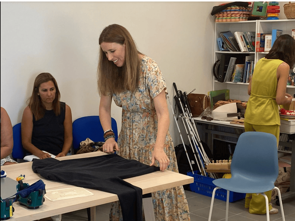 Há várias mulheres numa sala. Uma delas está de pé, a sorrir, e a dobrar uma peça de roupa escura em cima da mesa. Duas estão sentadas perto dela, a ver ou a ajudar. Outra mulher está ao fundo, junto a um balcão, a mexer em materiais. Na sala há livros, cestos, bengalas dobradas e coisas de trabalhos manuais. 