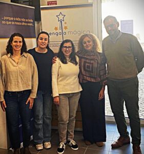 Cinco adultos estão juntos, sorridentes, em frente a dois painéis verticais. Um dos painéis tem a frase: "...porque a visão não se limita ao que os nossos olhos veem...". O outro representa a associação Bengala Mágica, dedicada ao apoio de pessoas com deficiência visual e crianças com necessidades educativas especiais. O ambiente transmite união e compromisso com a inclusão.