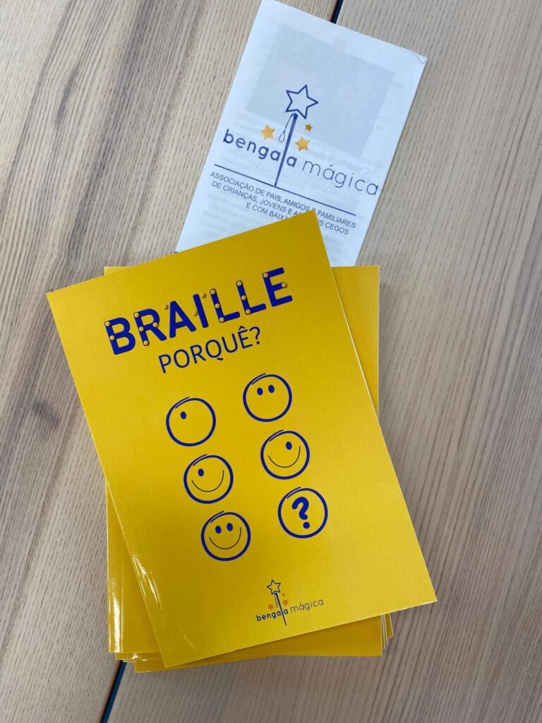 Sobre uma mesa de madeira está uma pequena pilha de livros com capa amarela. O título na capa é “Braille, Porquê?” e há seis círculos desenhados, cinco com caras sorridentes e um com um ponto de interrogação. Ao lado dos livros encontra-se um folheto da associação Bengala Mágica, que tem o logótipo composto por uma estrela no topo de uma varinha. Não aparecem pessoas na imagem, apenas os materiais impressos.