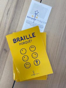 Capa do livro amarelo “Braille, Porquê?” com vários ícones circulares sorridentes e um ponto de interrogação, colocada sobre uma mesa de madeira ao lado de um folheto da Associação Bengala Mágica.